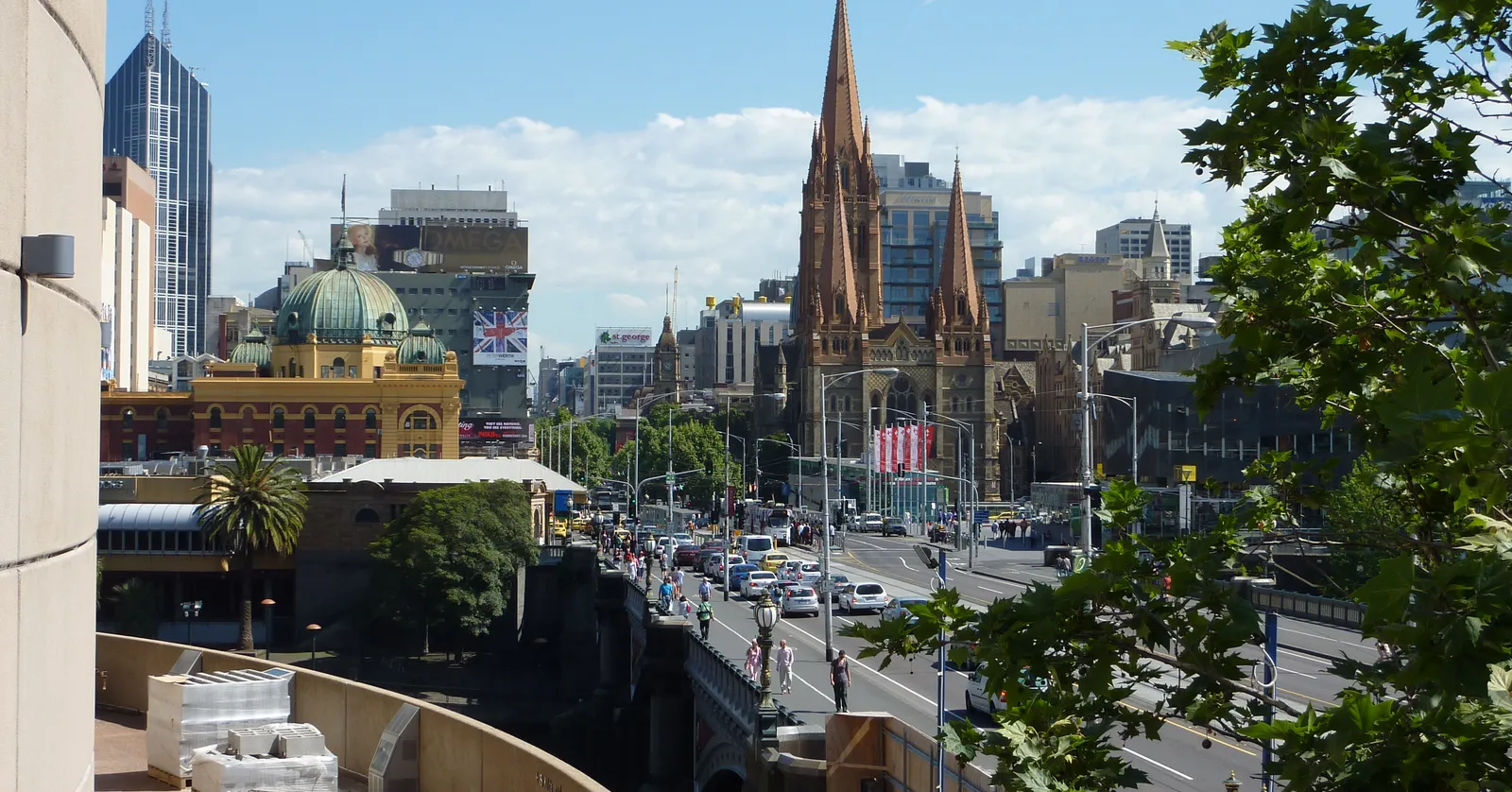 Looking down Swanston Street in Melbourne toward the green dome of Flinders Street Station on the left and the three sandstone spires of St Paul's Cathedral on the right, with Princes Bridge crossing the Yarra beyond