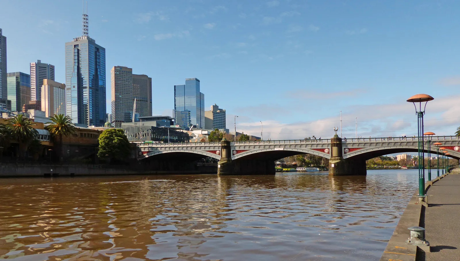 Princes Bridge crossing the Yarra River in Melbourne, its bluestone arches reflected in the brown water with the city towers rising behind on the north bank