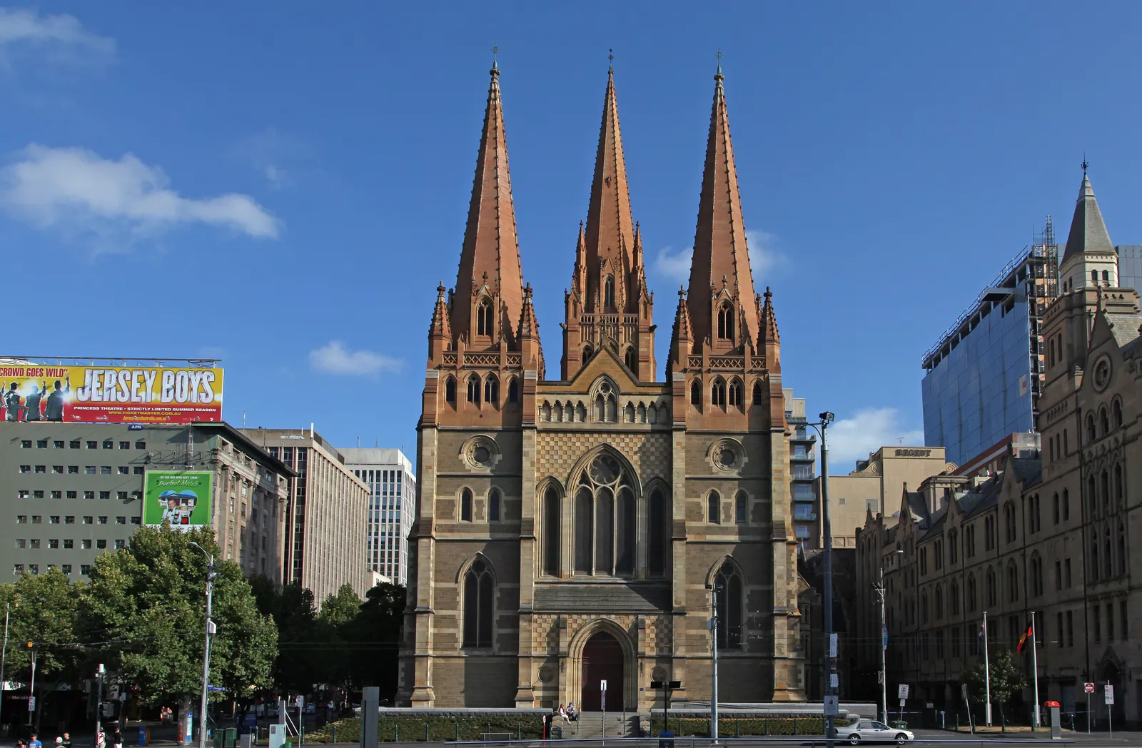 The west front of St Paul's Cathedral in Melbourne, three banded sandstone spires above a rose window and a recessed arched doorway, seen from the corner of Flinders and Swanston streets