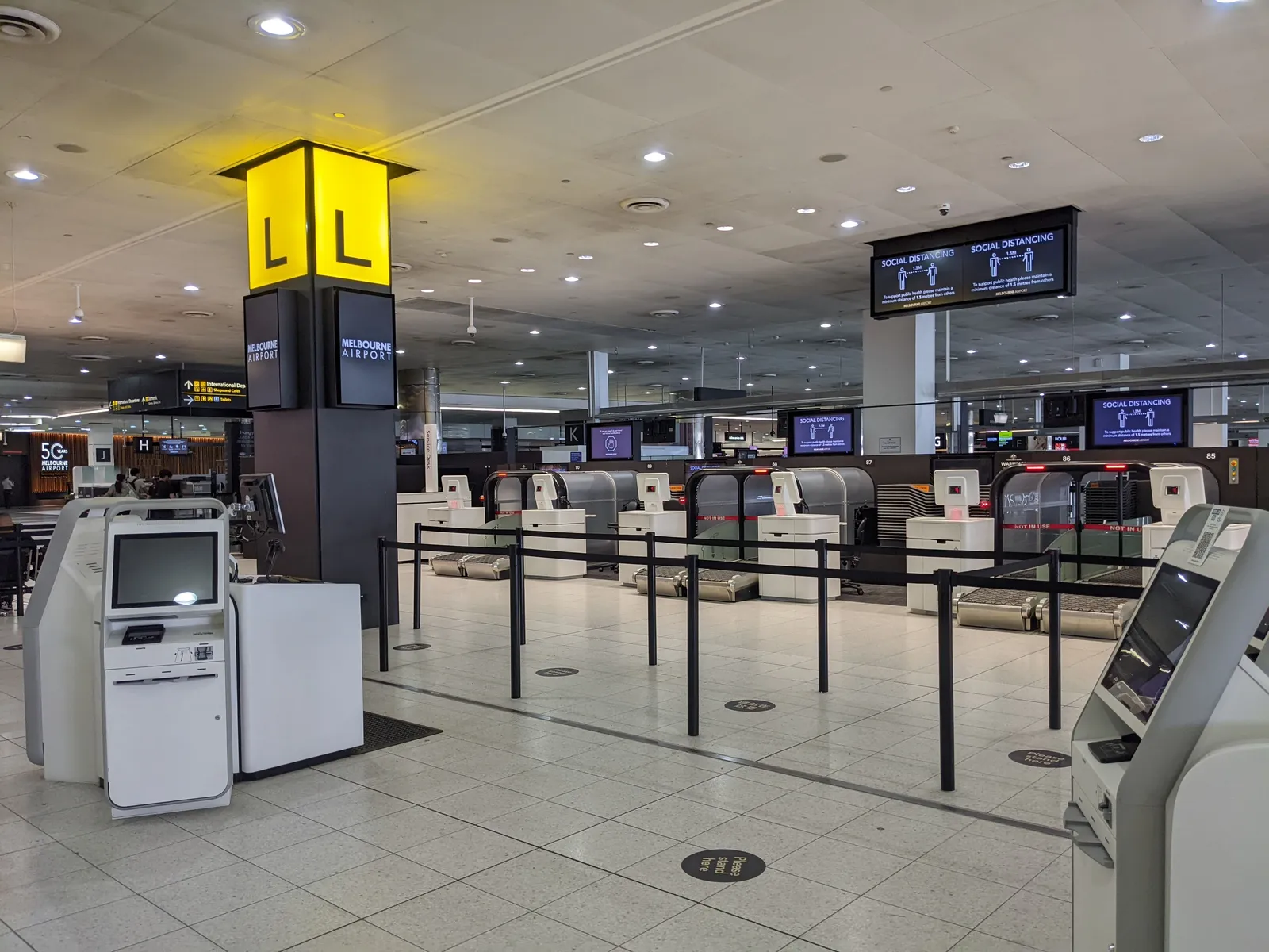 The check-in hall in Melbourne Airport's Terminal 2, with self-service bag drop machines behind roped lanes under a yellow aisle marker reading L