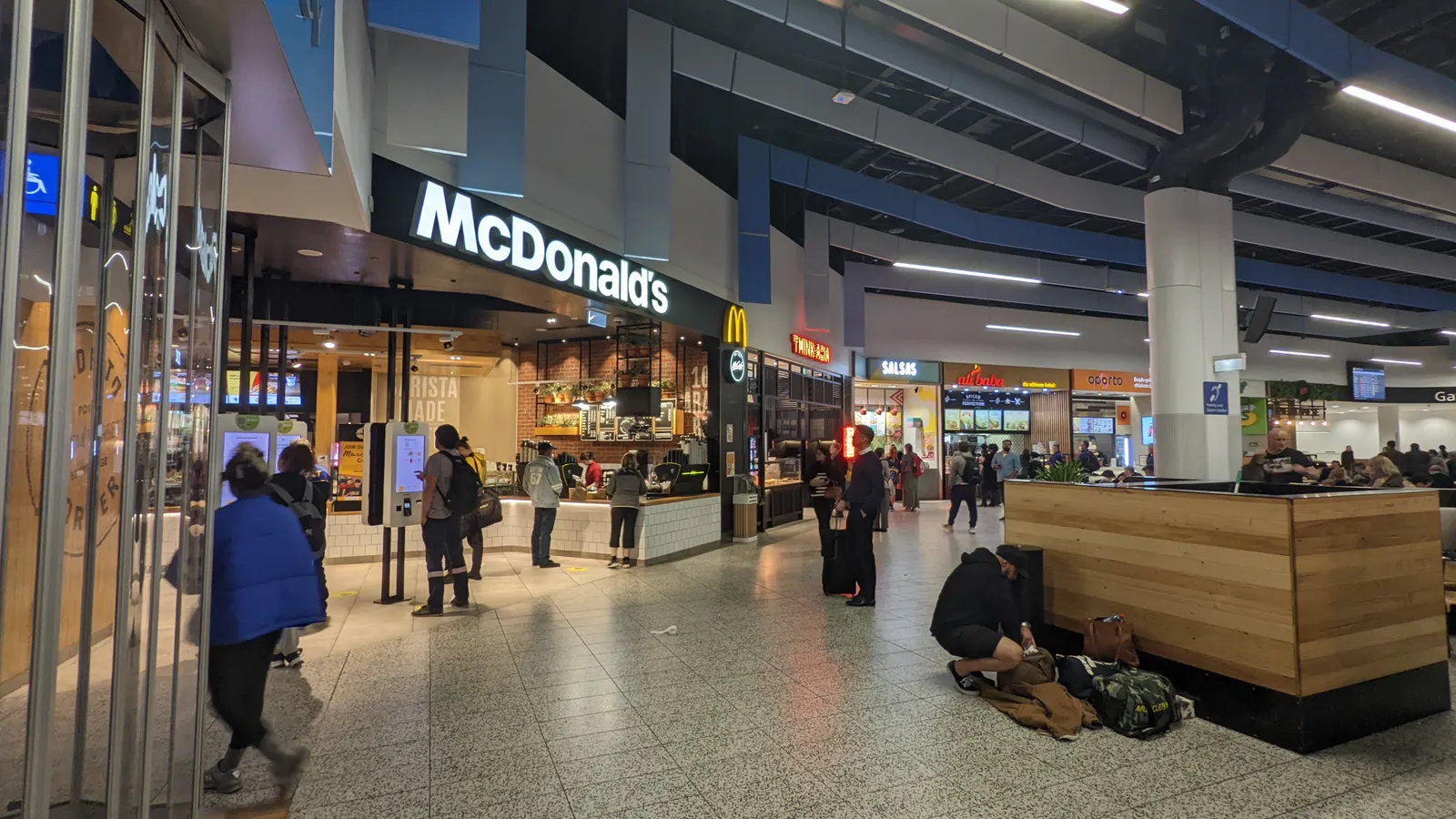 The concourse inside Melbourne Airport's Terminal 4, food outlets along one side and travellers sitting on a low timber bench with their bags