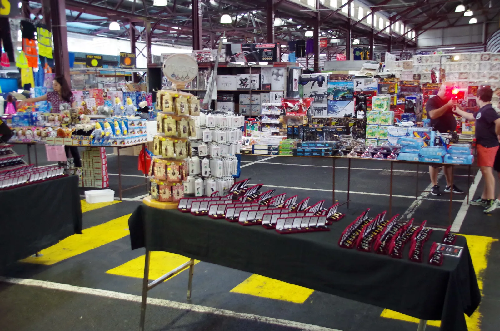 Trestle tables of goods under the open steel sheds at Queen Victoria Market in Melbourne, with narrow yellow-marked aisles running between the stalls