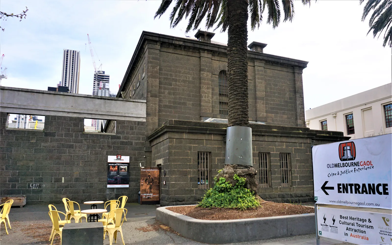 The bluestone walls and barred windows of Old Melbourne Gaol seen from its Russell Street forecourt, with a palm tree and an entrance sign in front