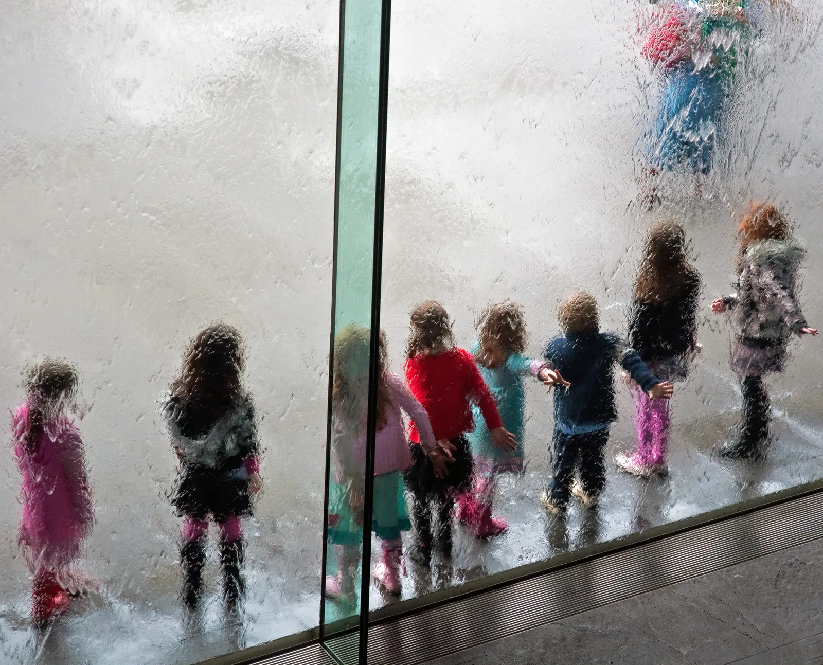 Children standing against the water wall at the entrance to the National Gallery of Victoria in Melbourne, their outlines blurred by the sheet of water running down the glass