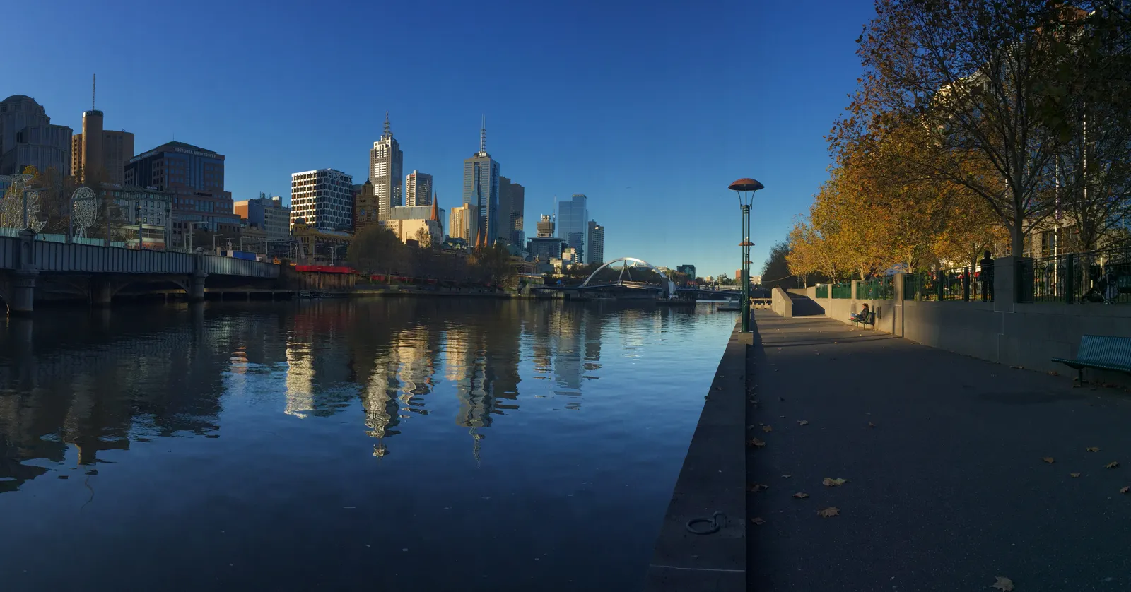 The Southbank promenade in Melbourne at low sun, a paved riverside walkway with autumn trees on the right and the city towers reflected in the still water of the Yarra