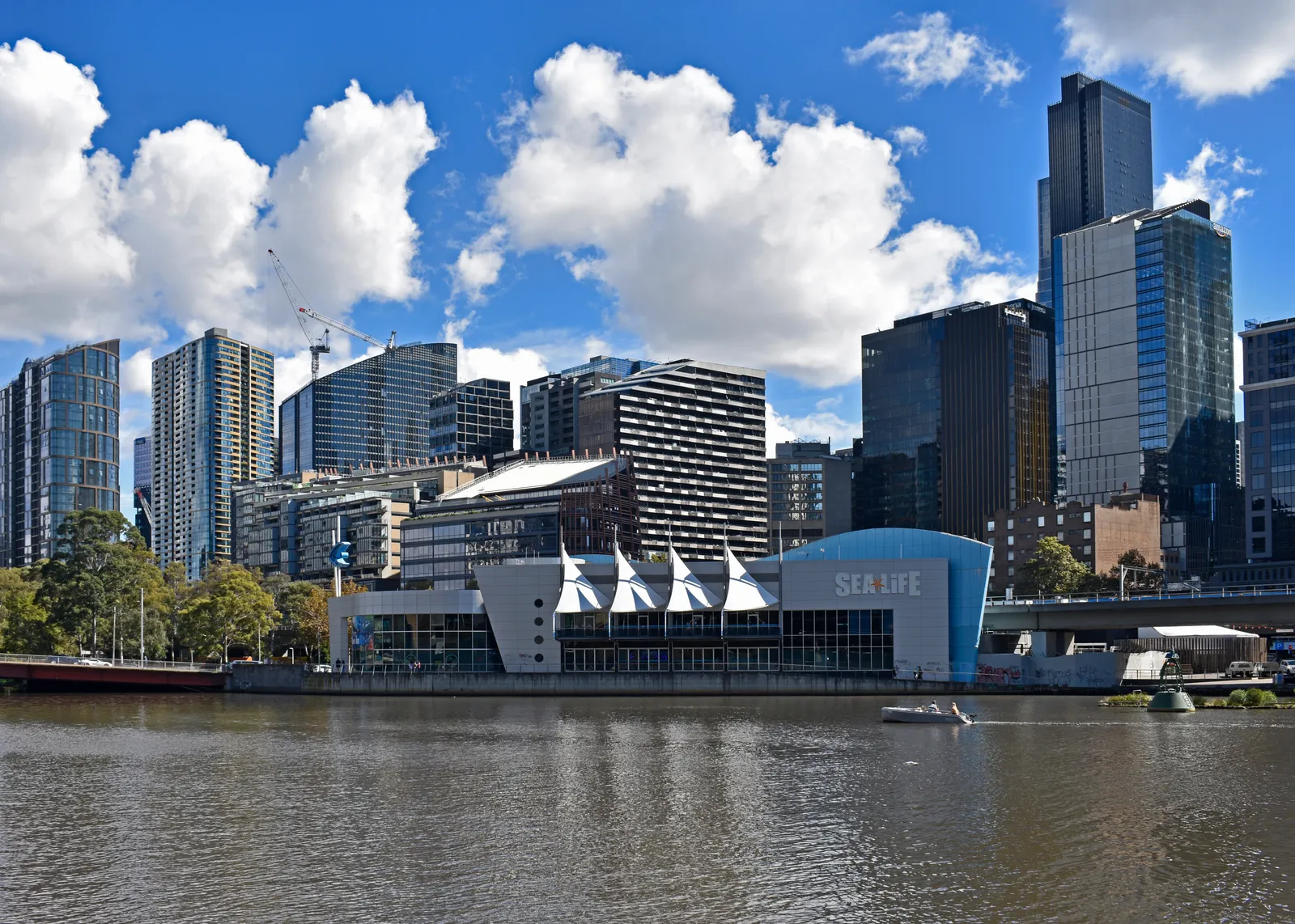 SEA LIFE Melbourne Aquarium seen across the Yarra River, a low blue building with white sail-shaped canopies along the north bank below the city towers