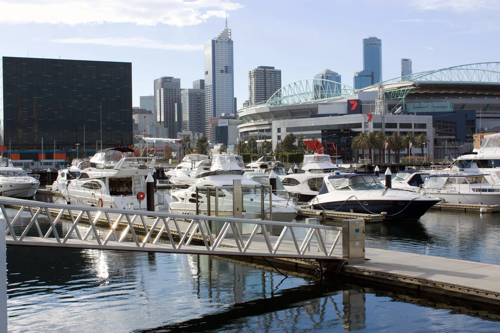 Boats moored along a floating pontoon at Victoria Harbour in Melbourne's Docklands, with the arched roof of Marvel Stadium and the city towers behind