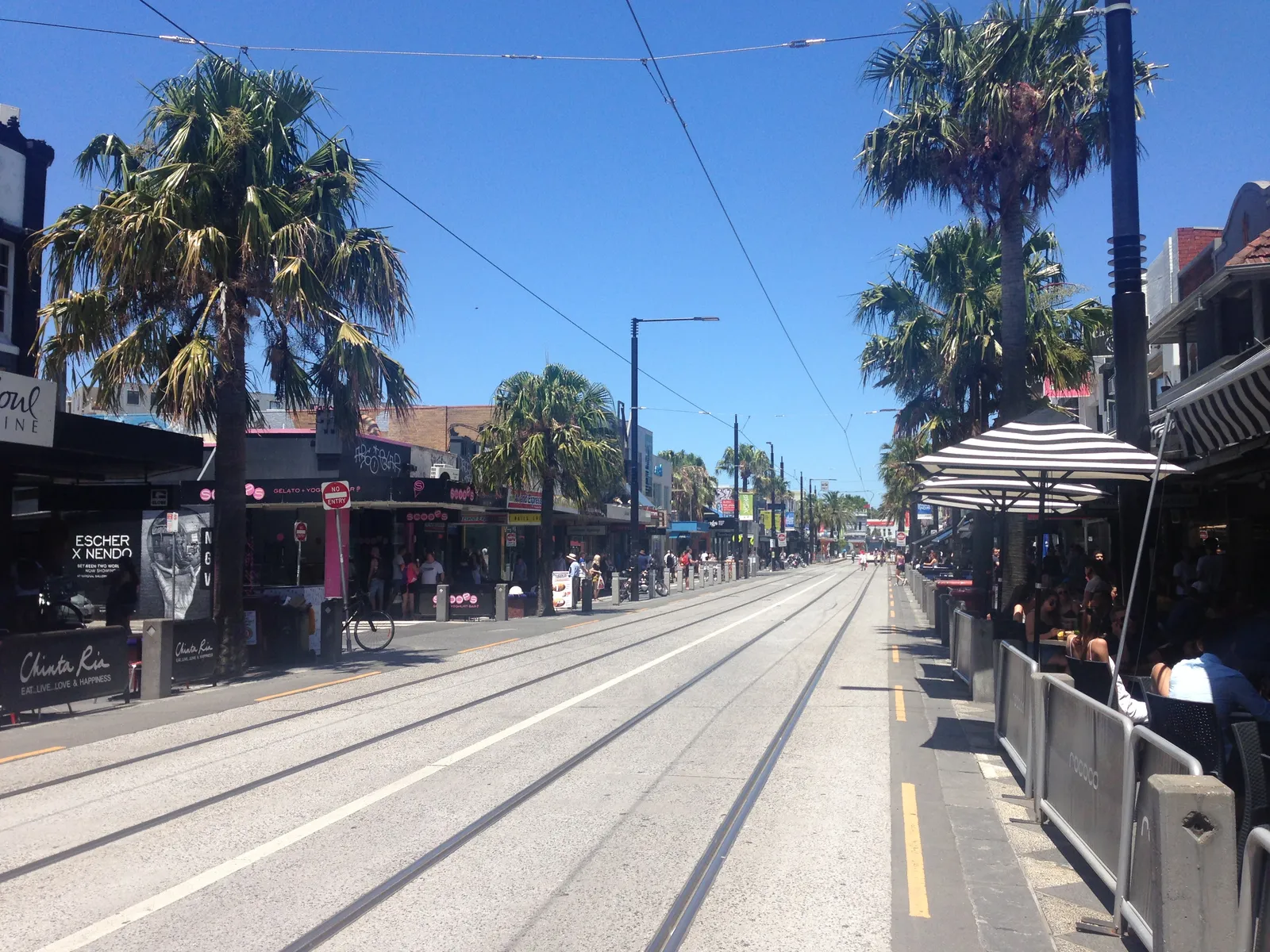 Acland Street in St Kilda, tram tracks running down the middle between palm trees, shopfront awnings and pavement tables on both sides
