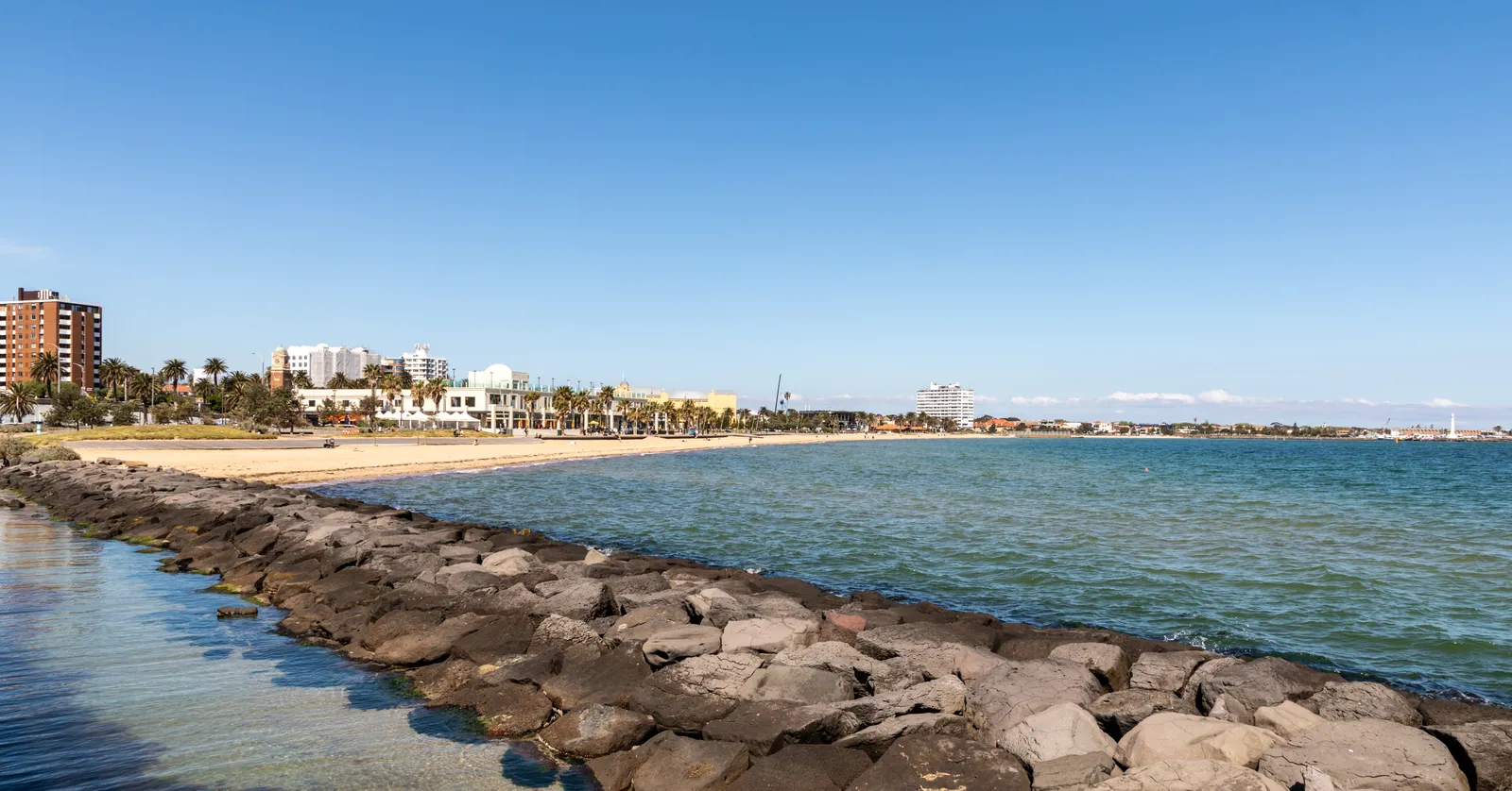 St Kilda Beach in Melbourne seen along the rock breakwater, with the curve of sand, palm trees and low white foreshore buildings behind it under a clear sky