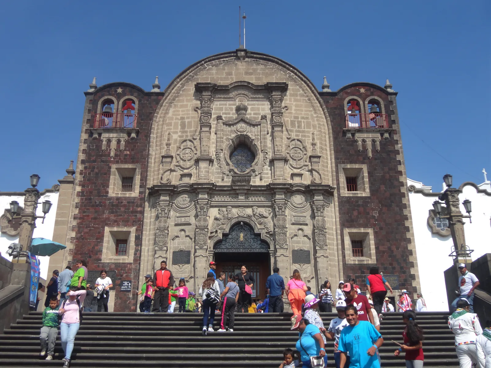 The Capilla del Cerrito at the top of Tepeyac hill in Mexico City, a small dark stone chapel with a carved doorway, reached by a broad flight of steps crowded with visitors