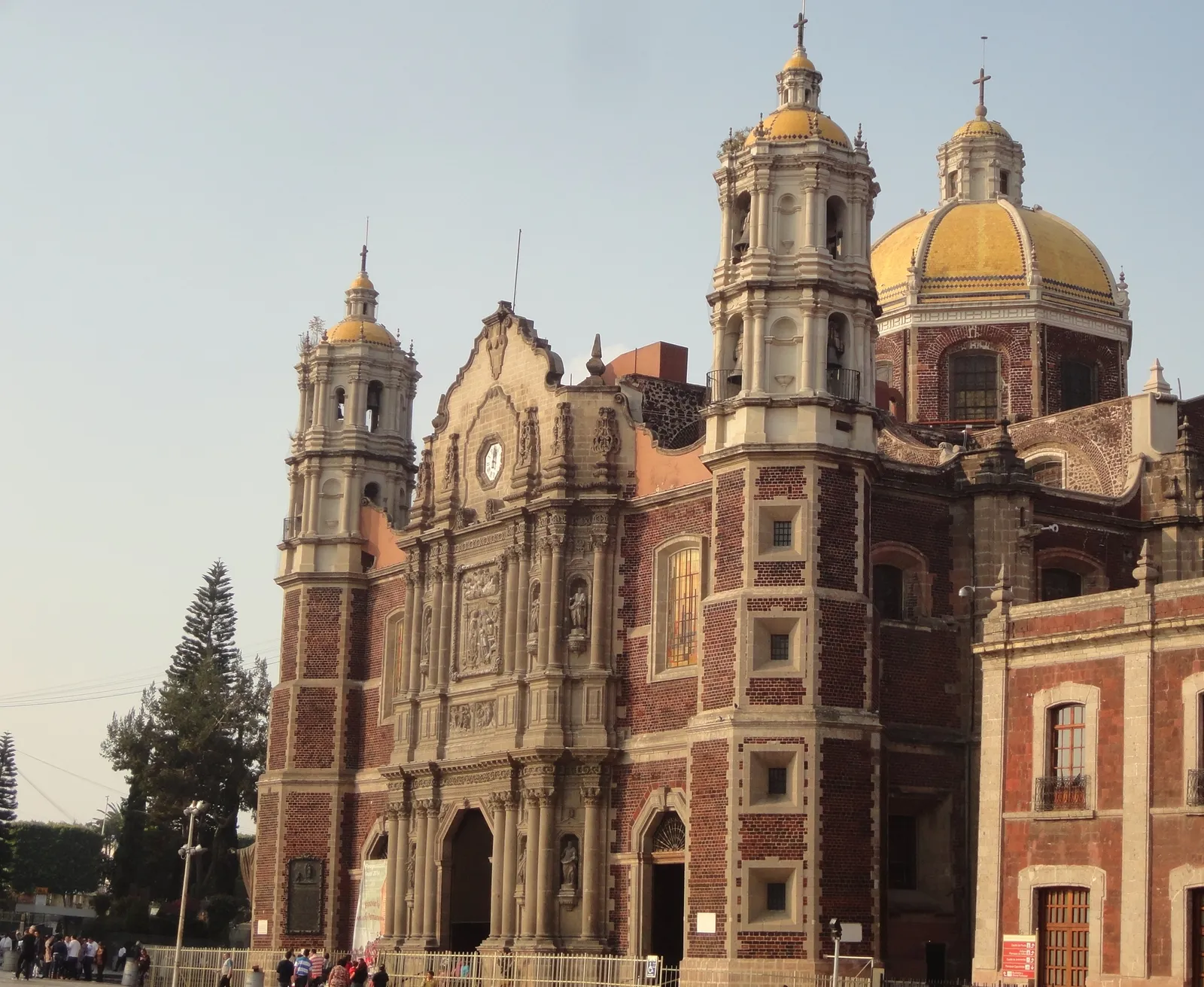The baroque front of the old Basilica of Guadalupe in Mexico City, in red volcanic stone with carved pale trim, twin bell towers and a yellow tiled dome behind
