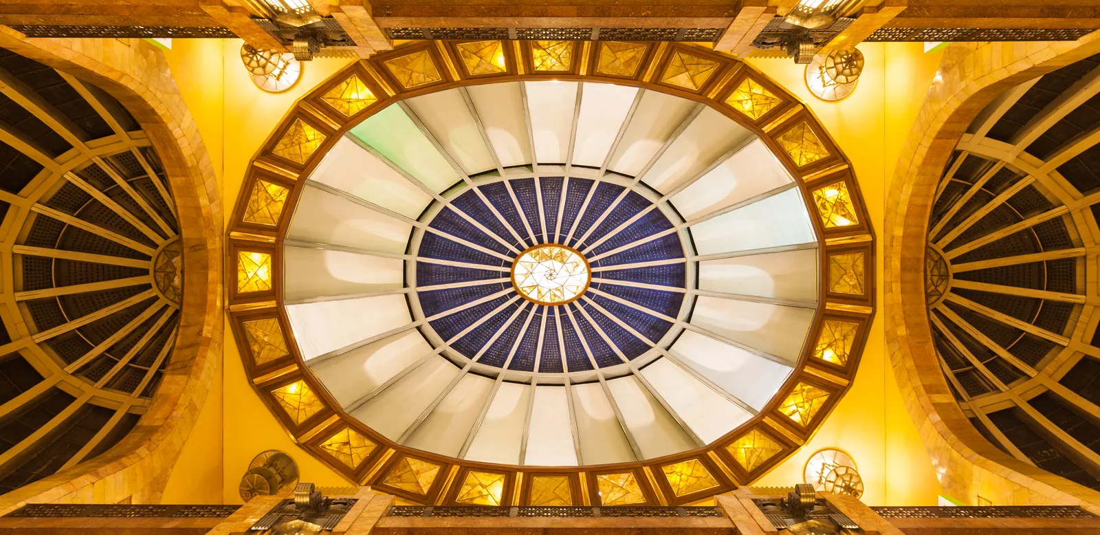 The oval art deco skylight over the lobby of the Palacio de Bellas Artes in Mexico City, ringed with amber glass panels and hanging lamps against yellow marble walls
