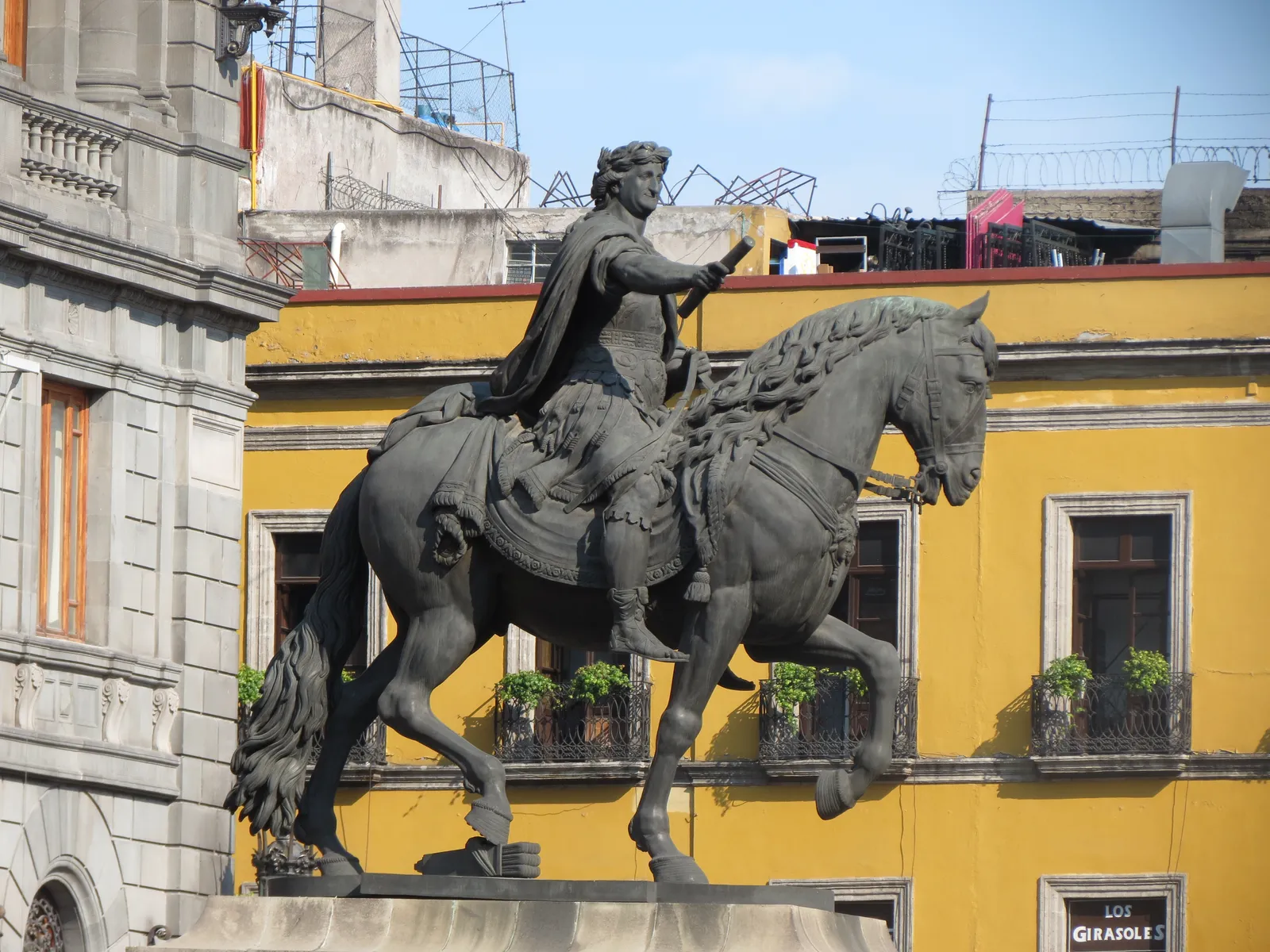 The bronze equestrian statue of Carlos IV known as El Caballito on Plaza Manuel Tolsá in Mexico City, the horse mid-stride against a yellow building behind it