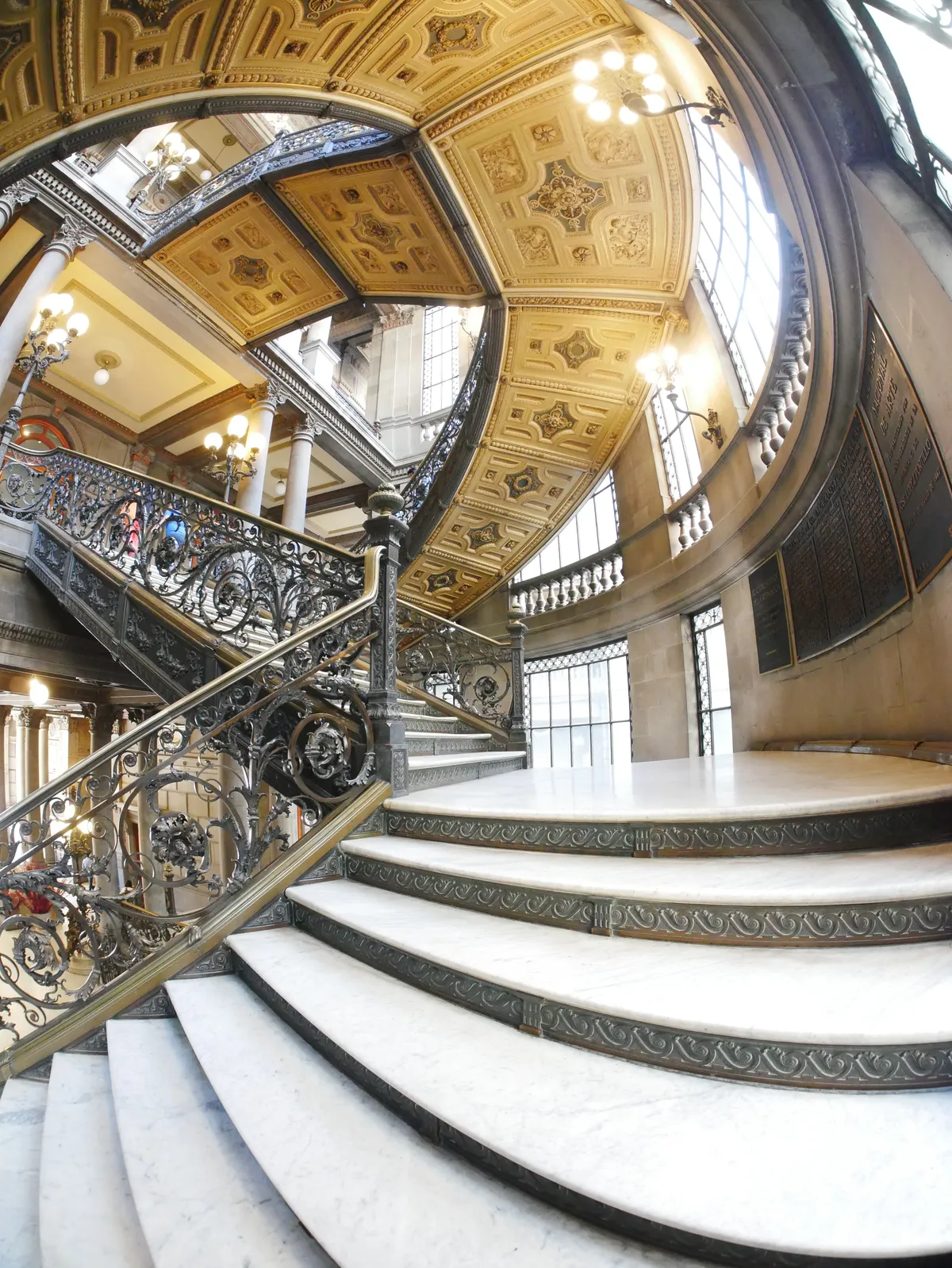 The grand marble staircase of the Museo Nacional de Arte in Mexico City, curving up under a gilded coffered ceiling behind ornate wrought-iron balustrades