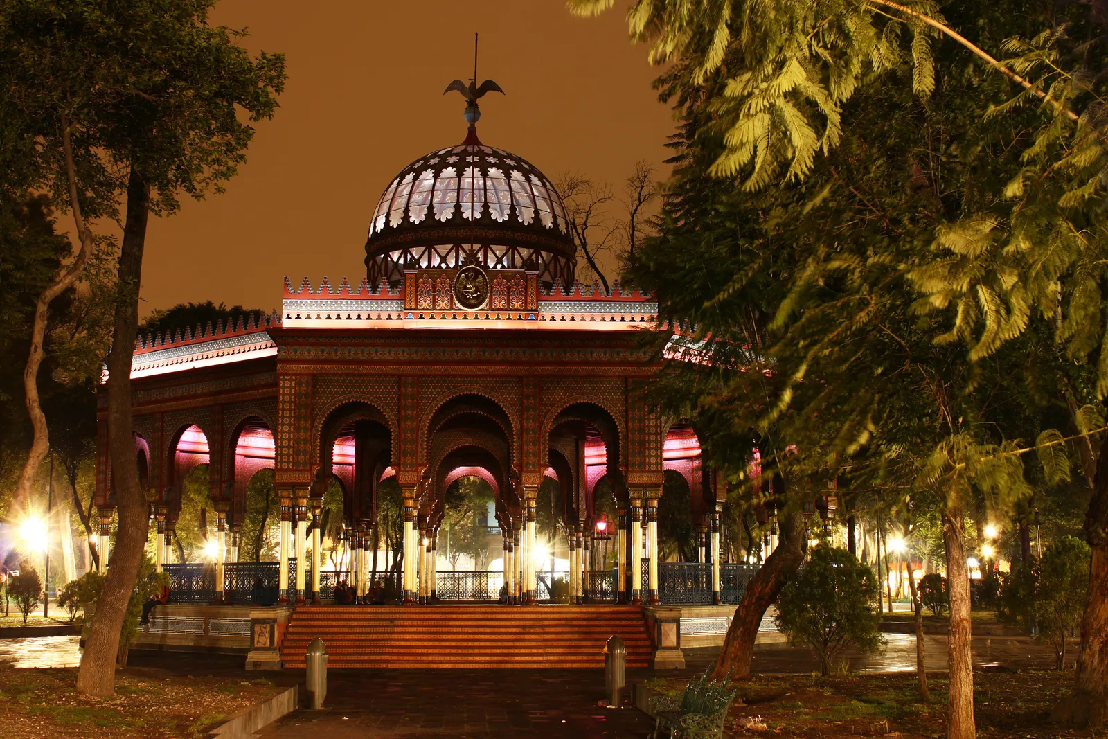 The Kiosco Morisco lit at night in the Alameda de Santa María la Ribera, a wrought-iron pavilion with horseshoe arches and a glazed openwork dome topped by an eagle