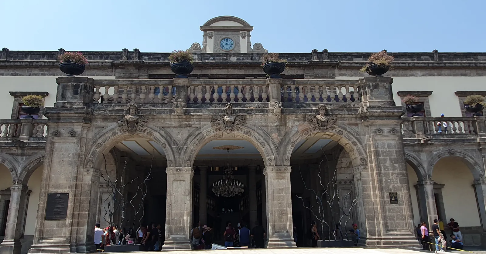 The arcaded main front of Chapultepec Castle in Mexico City, with a clock tower above the parapet and visitors standing under its three stone arches
