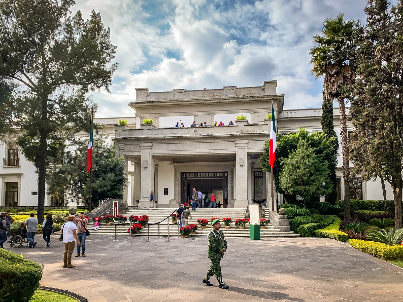 The white colonnaded front of the former presidential residence at Los Pinos in Mexico City, with Mexican flags either side of the entrance and visitors crossing the forecourt