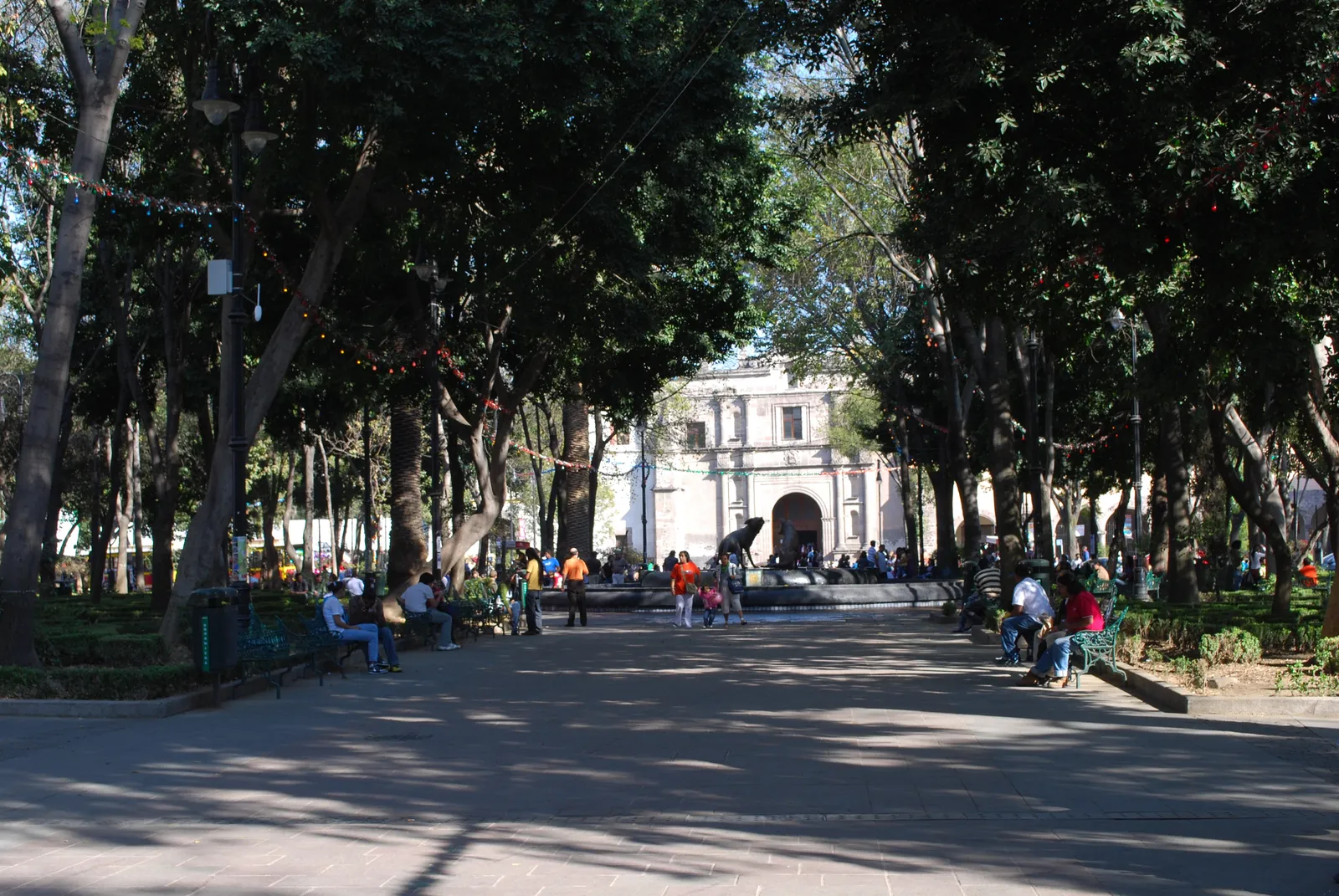 The tree-shaded Jardín Centenario in Coyoacán, Mexico City, with people on benches along a broad paved walk and the coyote fountain and church arch at the far end