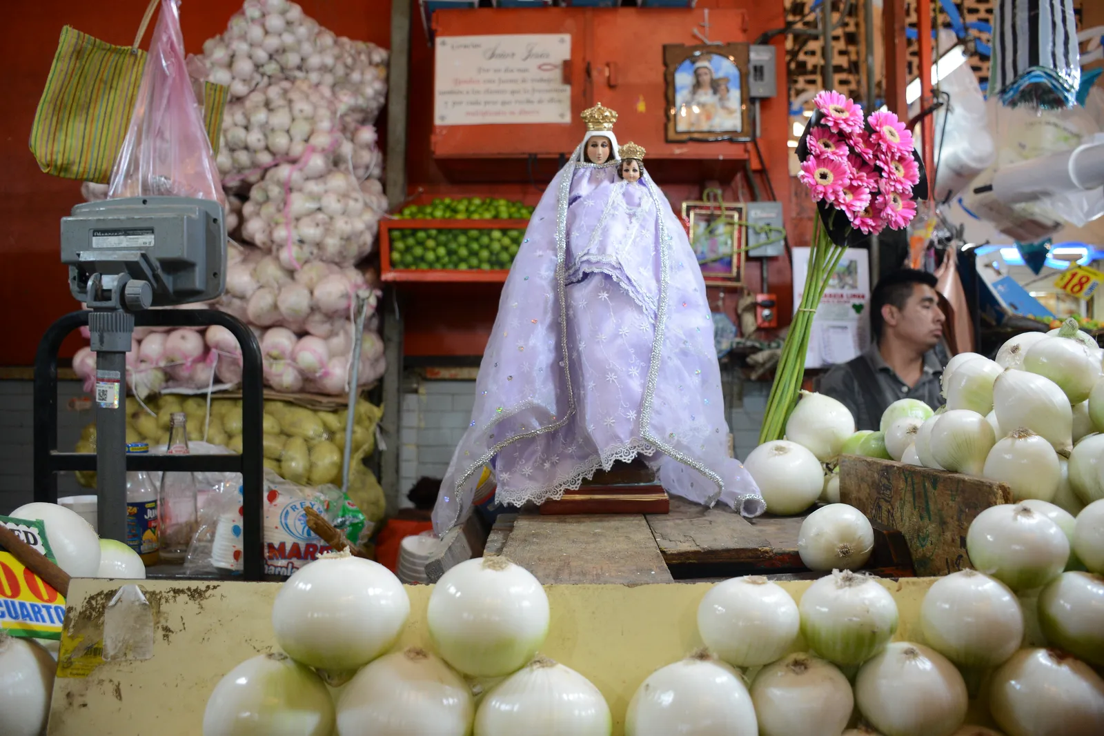 A produce stall inside the Mercado de La Merced in Mexico City, onions and garlic stacked around a robed religious figure with a stallholder behind the counter