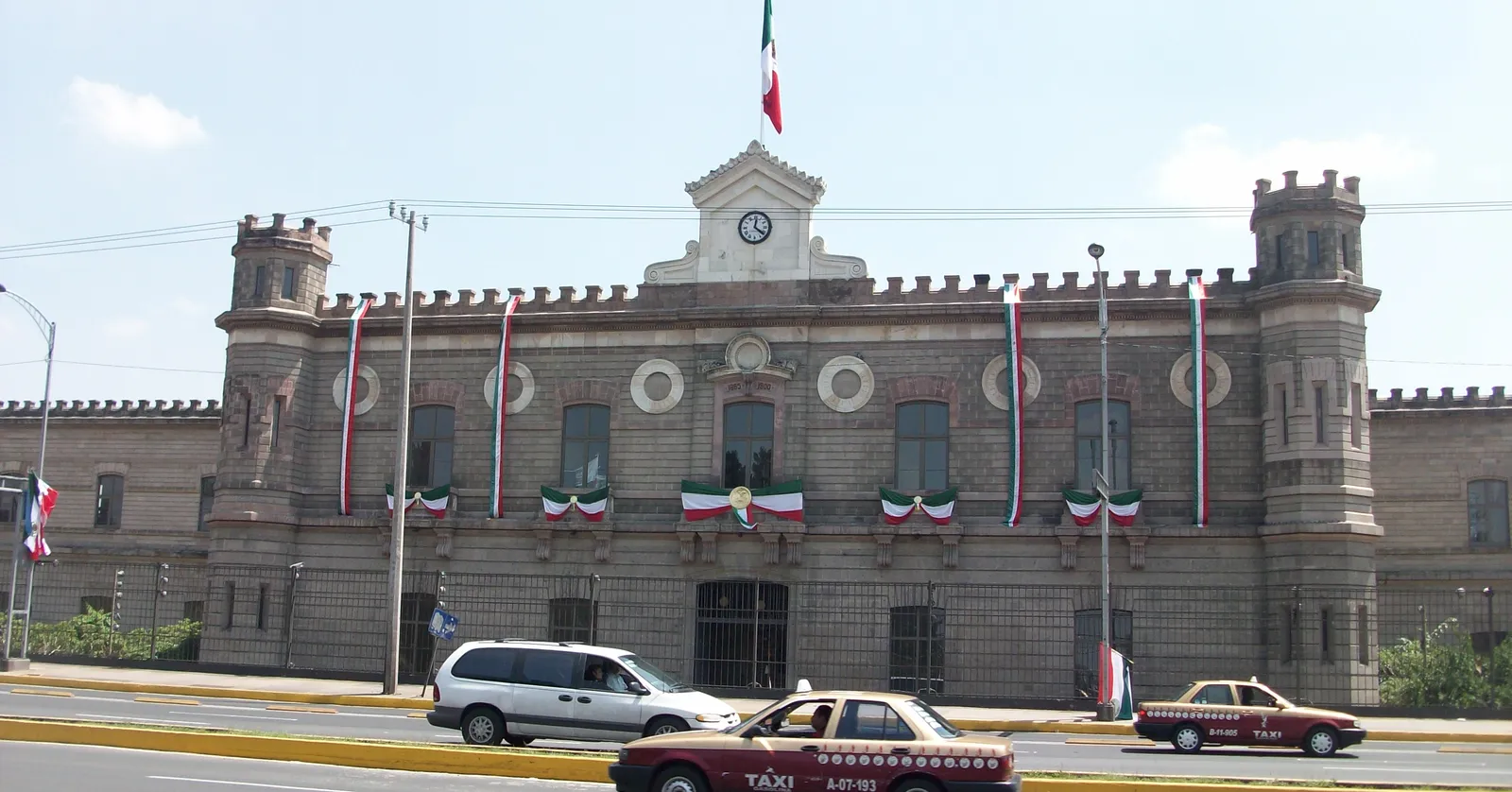 The castellated grey stone front of the Palacio de Lecumberri in Mexico City, with a clock tower and flagpole above the entrance and traffic passing on the avenue in front