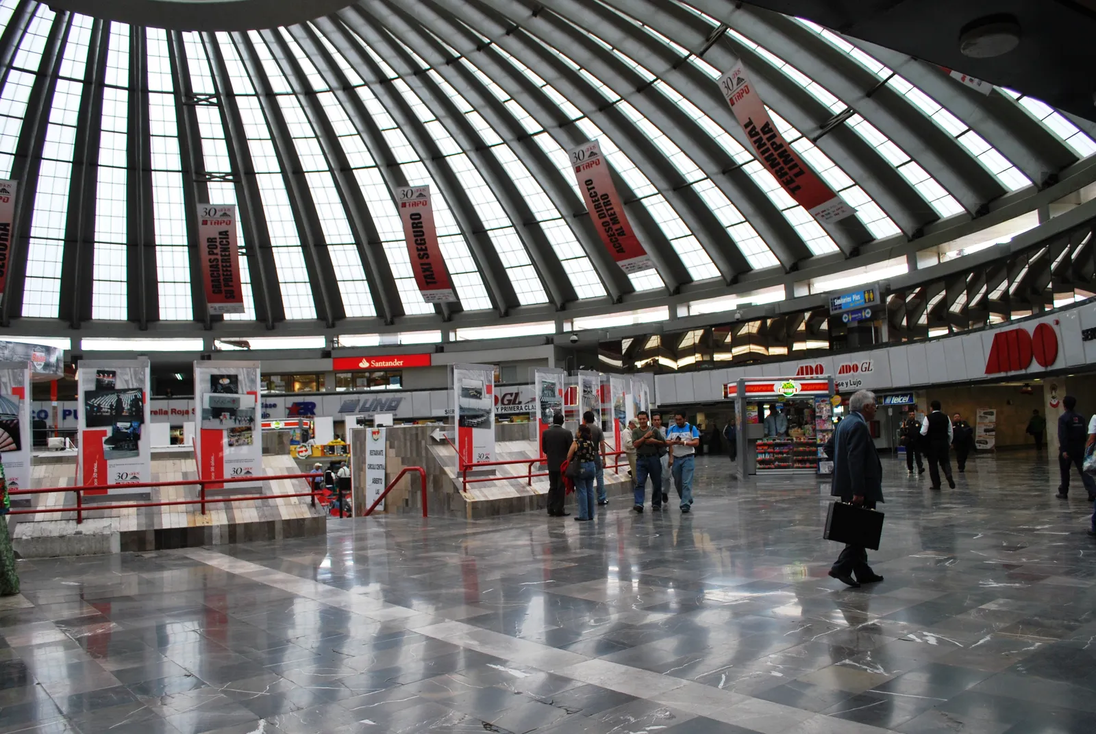 The circular concourse of the TAPO bus terminal in Mexico City under its ribbed glazed dome, with ticket counters around the edge and passengers crossing the polished floor