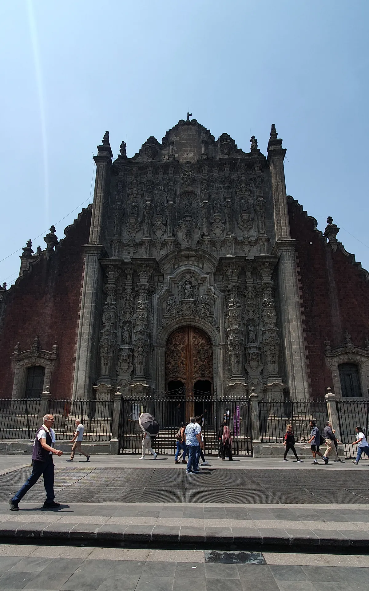 The east front of the Sagrario Metropolitano beside Mexico City's cathedral, a densely carved pale stone facade set into dark red volcanic walls, with people passing on the pavement