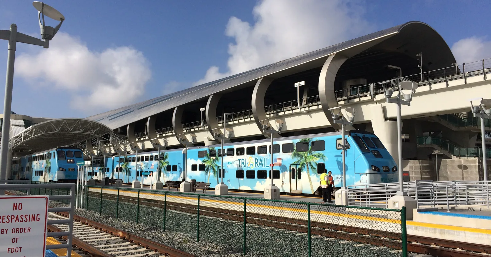 A blue Tri-Rail commuter train painted with palm trees at the Miami Airport station, under the station's curved white canopy