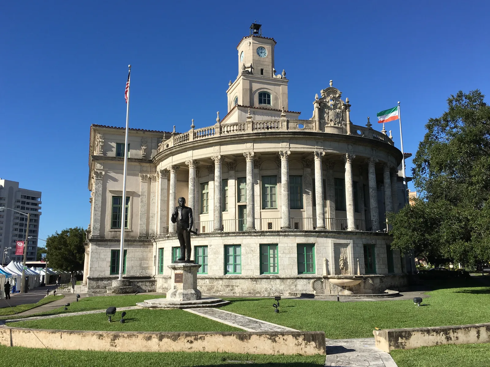 Coral Gables City Hall in Miami, a limestone Mediterranean Revival building with a curved colonnade and a clock tower on top, a statue on a pedestal and a lawn in front