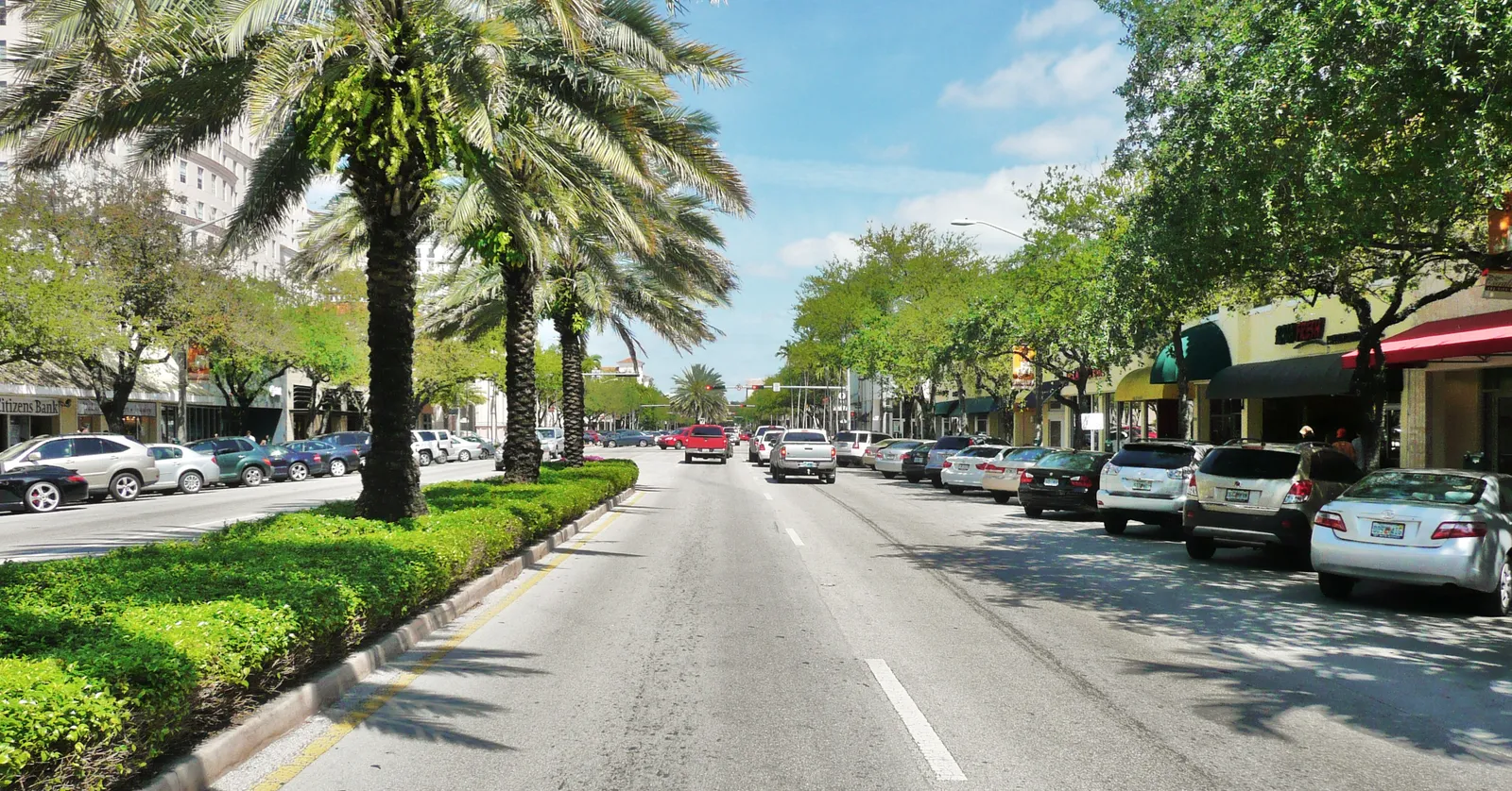 Miracle Mile in Coral Gables, Miami, a wide stretch of Coral Way with a planted median of palms and hedges, cars parked along both sides and low shopfronts under awnings