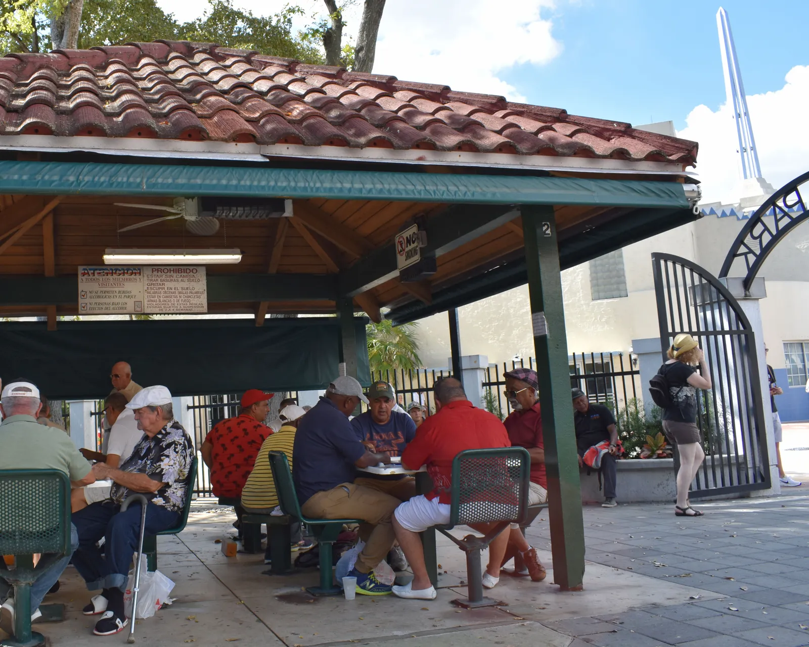 Men playing dominoes at tables under the tiled pavilion of Domino Park on Calle Ocho in Little Havana, Miami, with the park's iron gate and a white spire behind