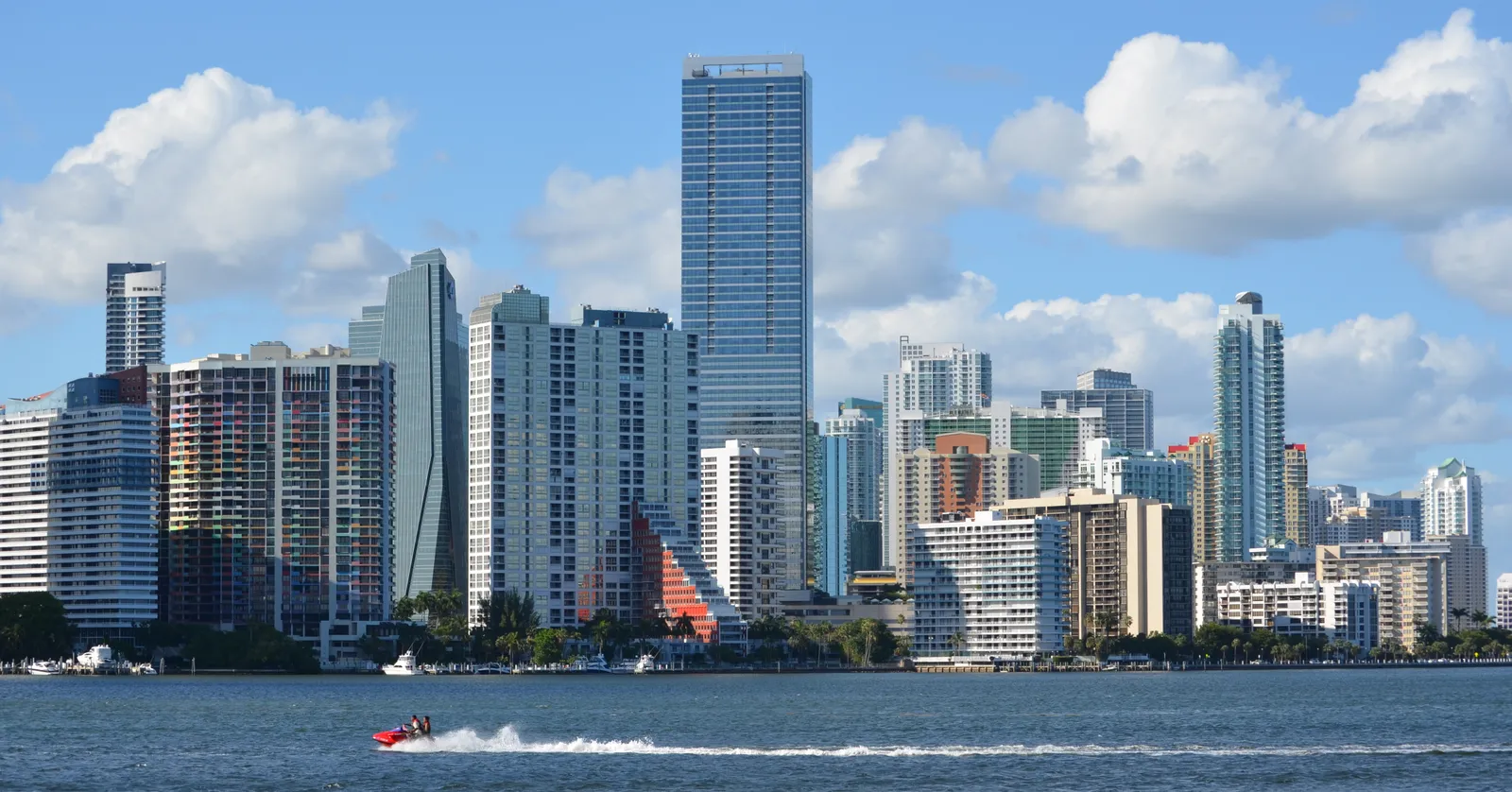 Brickell's skyline in Miami seen from Biscayne Bay, a dense row of glass and concrete towers along the waterfront under a blue sky with white clouds, a jet ski crossing the water in front