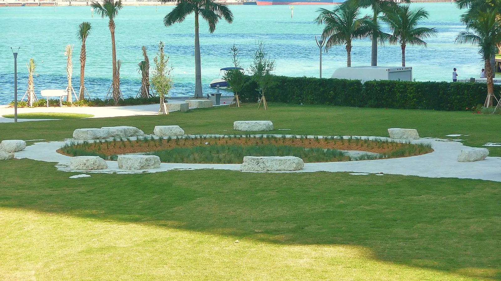 The Miami Circle park at Brickell Point in Miami, a ring of planted ground outlined by stone blocks on a lawn, with palm trees and the turquoise water of Biscayne Bay behind
