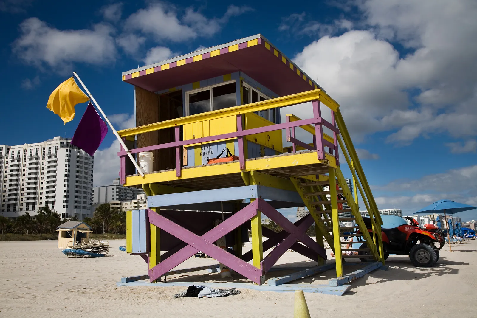 A yellow and pink lifeguard tower on the sand at Lummus Park beach in Miami Beach, with a cross-braced base, a flag flying beside it and beachfront towers behind