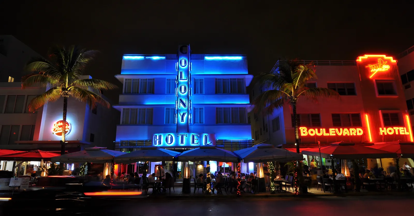 Ocean Drive on South Beach at dusk, the Colony Hotel's blue neon sign running down its Art Deco tower between palm trees, with the red neon of the Boulevard Hotel next door and café umbrellas along the sidewalk