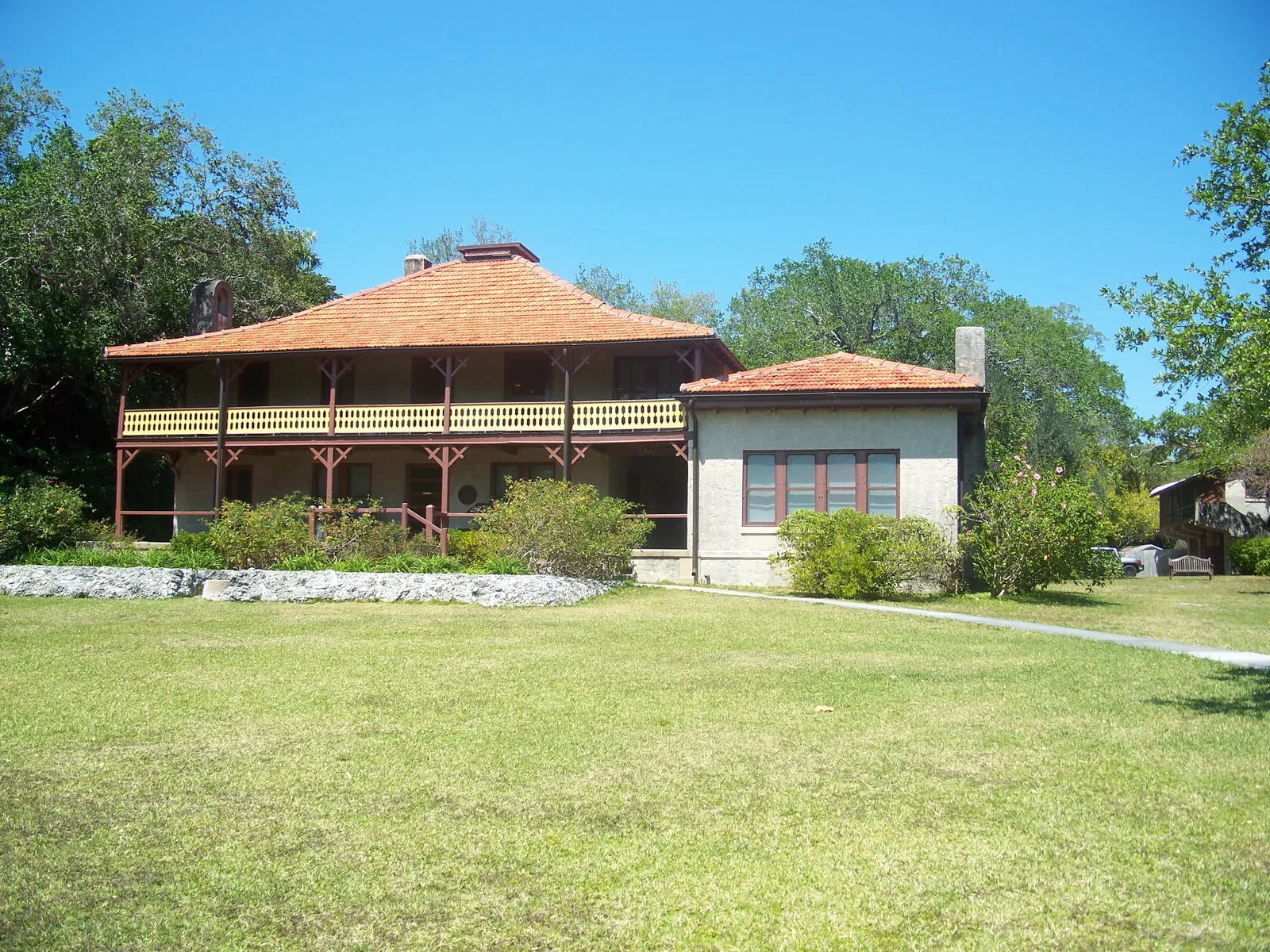 The Barnacle in Coconut Grove, Miami, a two-story wooden house with a red hip roof and a long upper veranda with a yellow railing, set back across a wide lawn