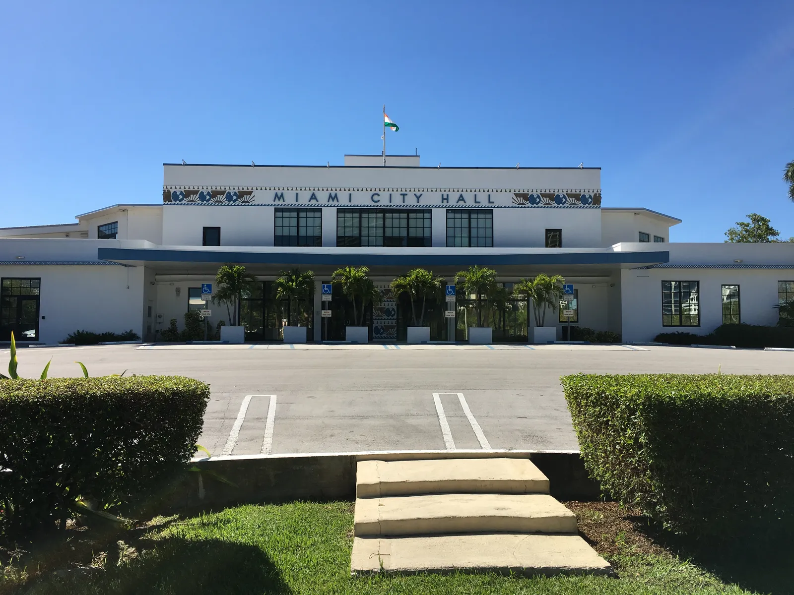 Miami City Hall at Dinner Key in Coconut Grove, a white 1930s terminal building with a decorated band below its roofline lettered Miami City Hall, palms along the front and a lawn and steps in the foreground