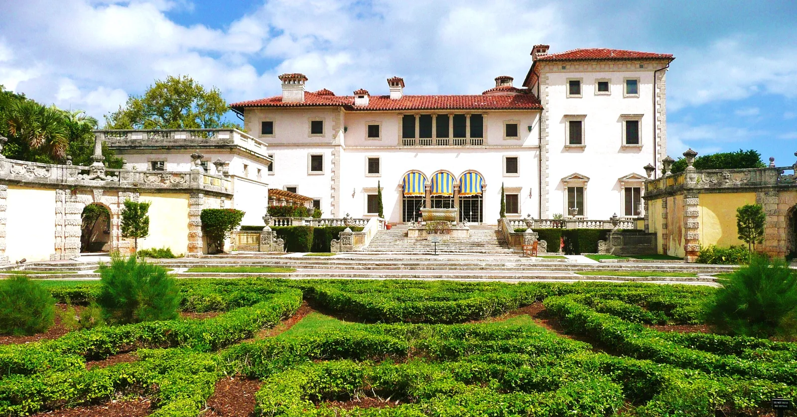 Vizcaya's main house in Miami, a cream Italian Renaissance-style villa with a red tile roof, seen across clipped hedges laid out in a pattern in the formal garden
