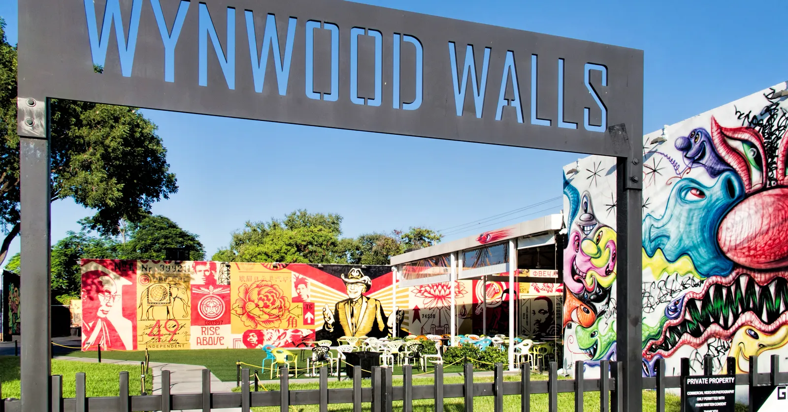 The Wynwood Walls sign in Miami, steel letters on a frame over the entrance gate, with brightly painted murals on the low warehouse buildings and a lawn behind it