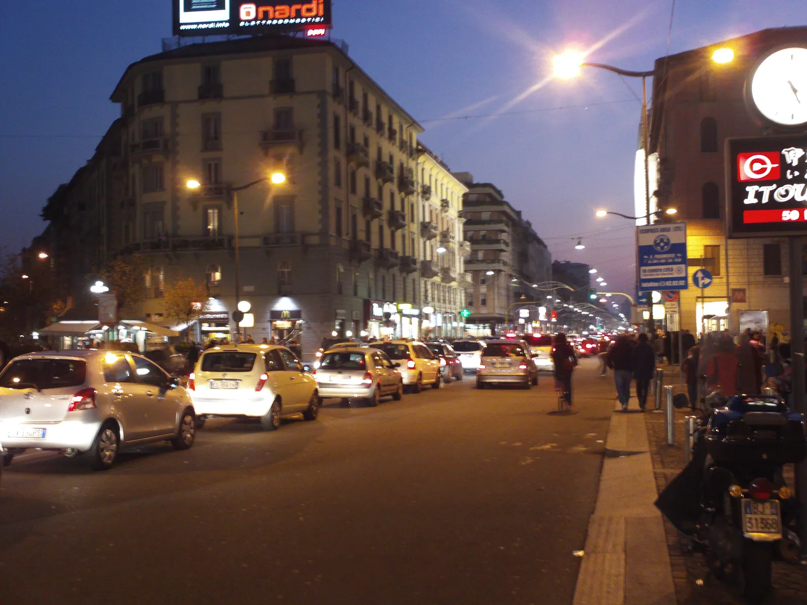 Corso Buenos Aires in Milan at dusk, a wide shopping avenue lined with storefronts, streetlights, and pedestrians
