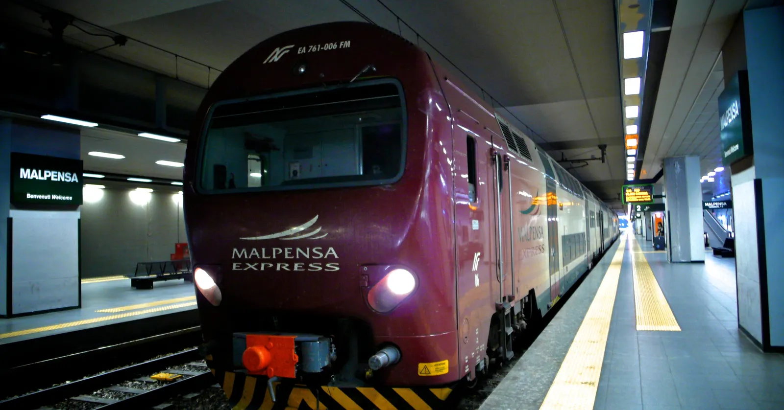 A burgundy Malpensa Express train waiting at the underground platform of Milan Malpensa airport station, with the Malpensa station sign on the left