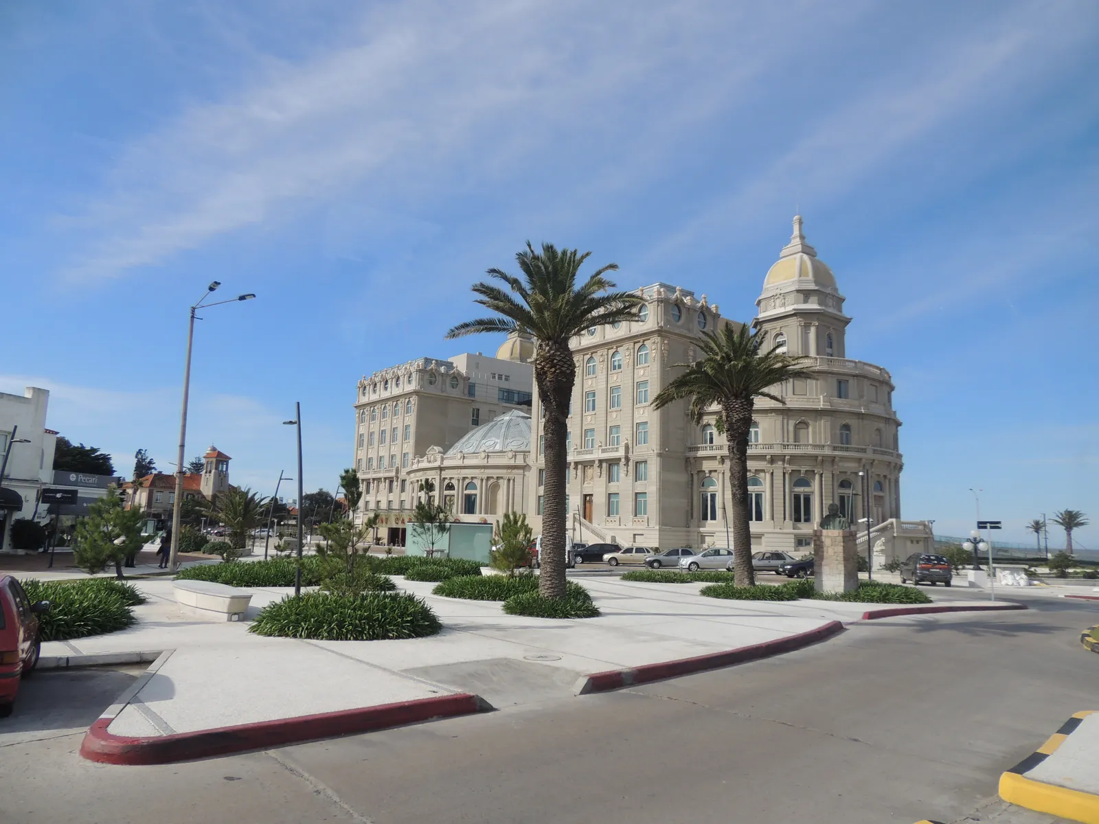 The Hotel Carrasco in Montevideo, a pale stone palace hotel with a domed corner tower and tall arched windows, standing behind palm trees at a wide seafront junction under a blue sky
