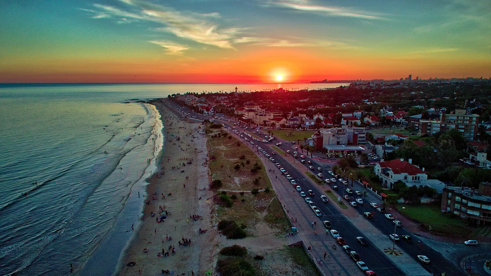An aerial view along Carrasco beach at the eastern edge of Montevideo at sunset, a long pale strand meeting the Río de la Plata on one side and the coast road and low houses of the neighbourhood on the other