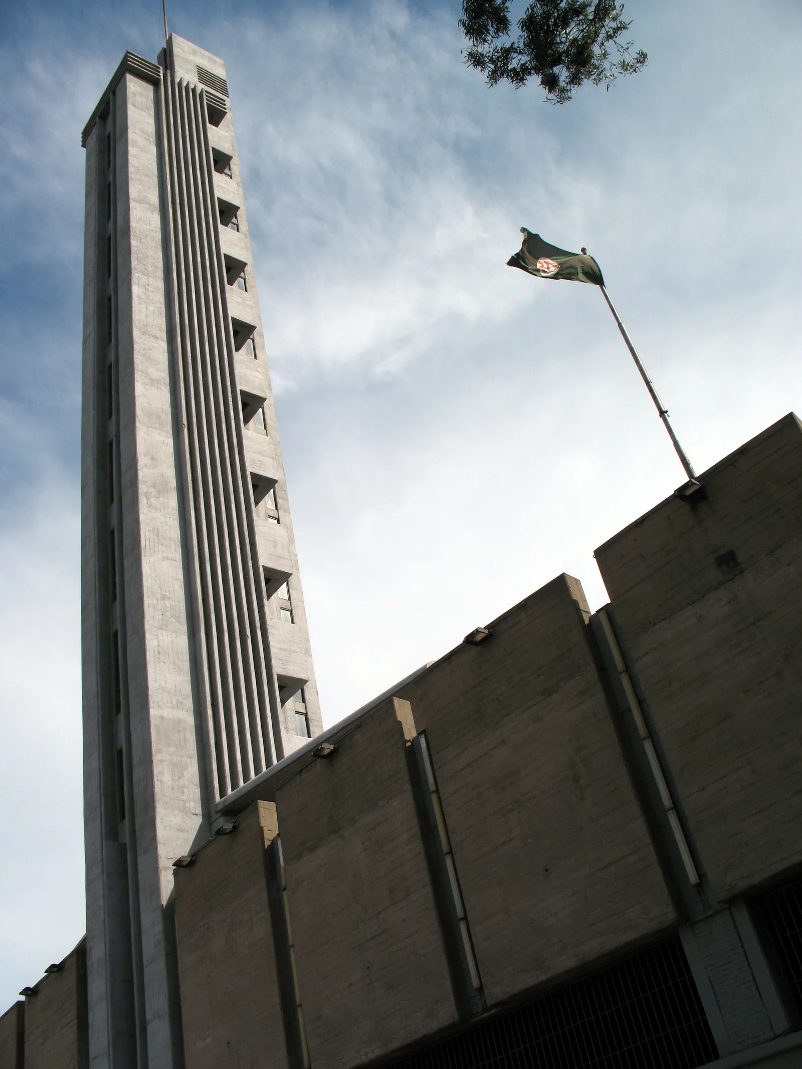 Looking up at the Torre de los Homenajes at Montevideo's Estadio Centenario, a tall fluted concrete tower against a partly cloudy sky, with a flag flying from the grandstand roof beside it