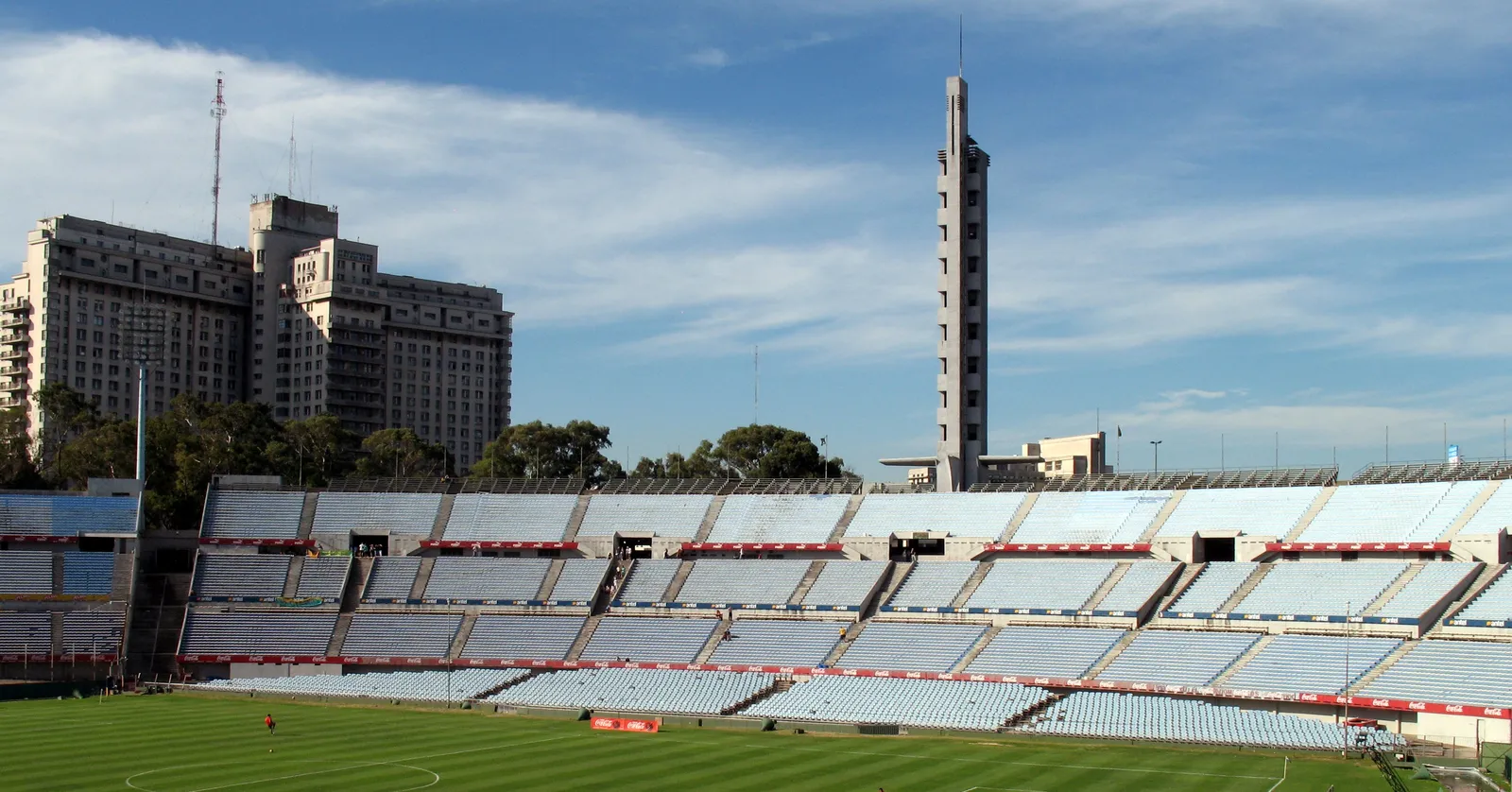 The Olímpica grandstand of Montevideo's Estadio Centenario, empty pale-blue seating tiers under the Torre de los Homenajes rising behind it, with an apartment building on the skyline beyond