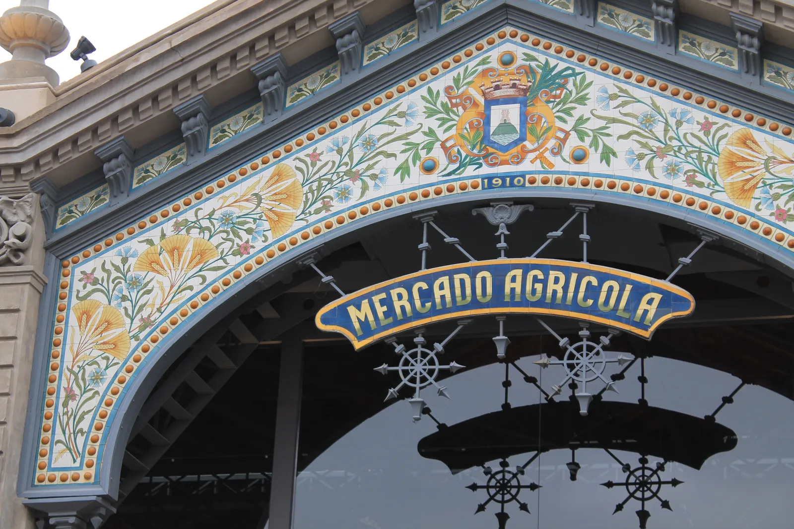The ornate tiled entrance arch of the Mercado Agrícola de Montevideo, painted flowers and a coat of arms around a scrolled sign reading Mercado Agricola with the date 1910 worked into the tilework above it