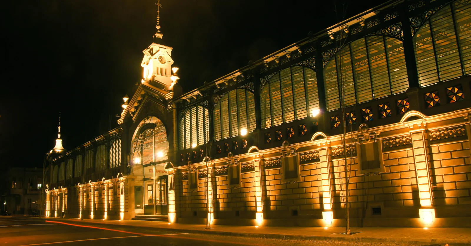 The illuminated iron-and-glass facade of the Mercado Agrícola de Montevideo at night, its central clock tower lit up above a long arcaded stone wall on an empty street