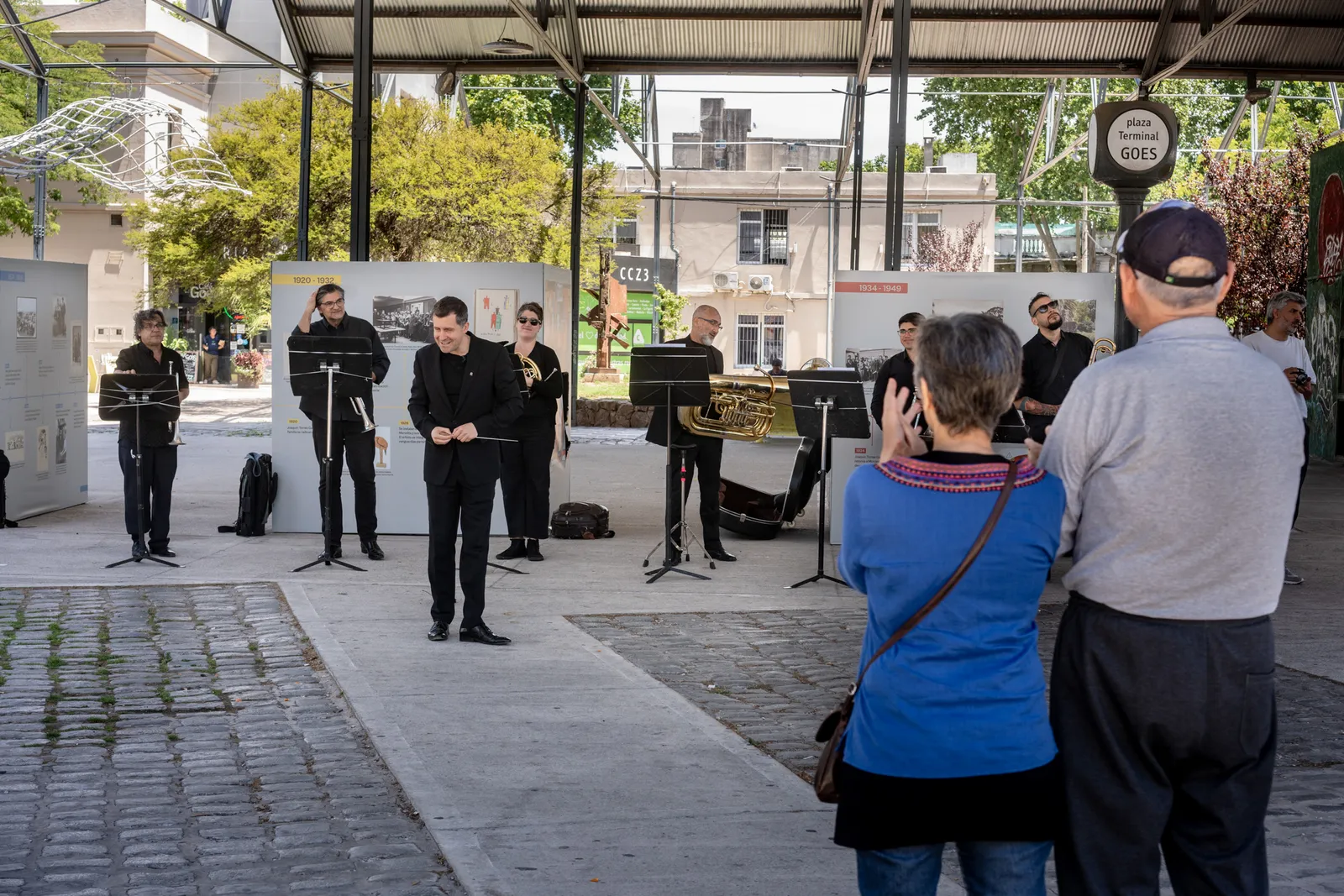 Musicians performing under the steel canopy of the old Terminal Goes in Montevideo, now a covered public plaza, with onlookers gathered around and a GOES sign visible on a nearby post
