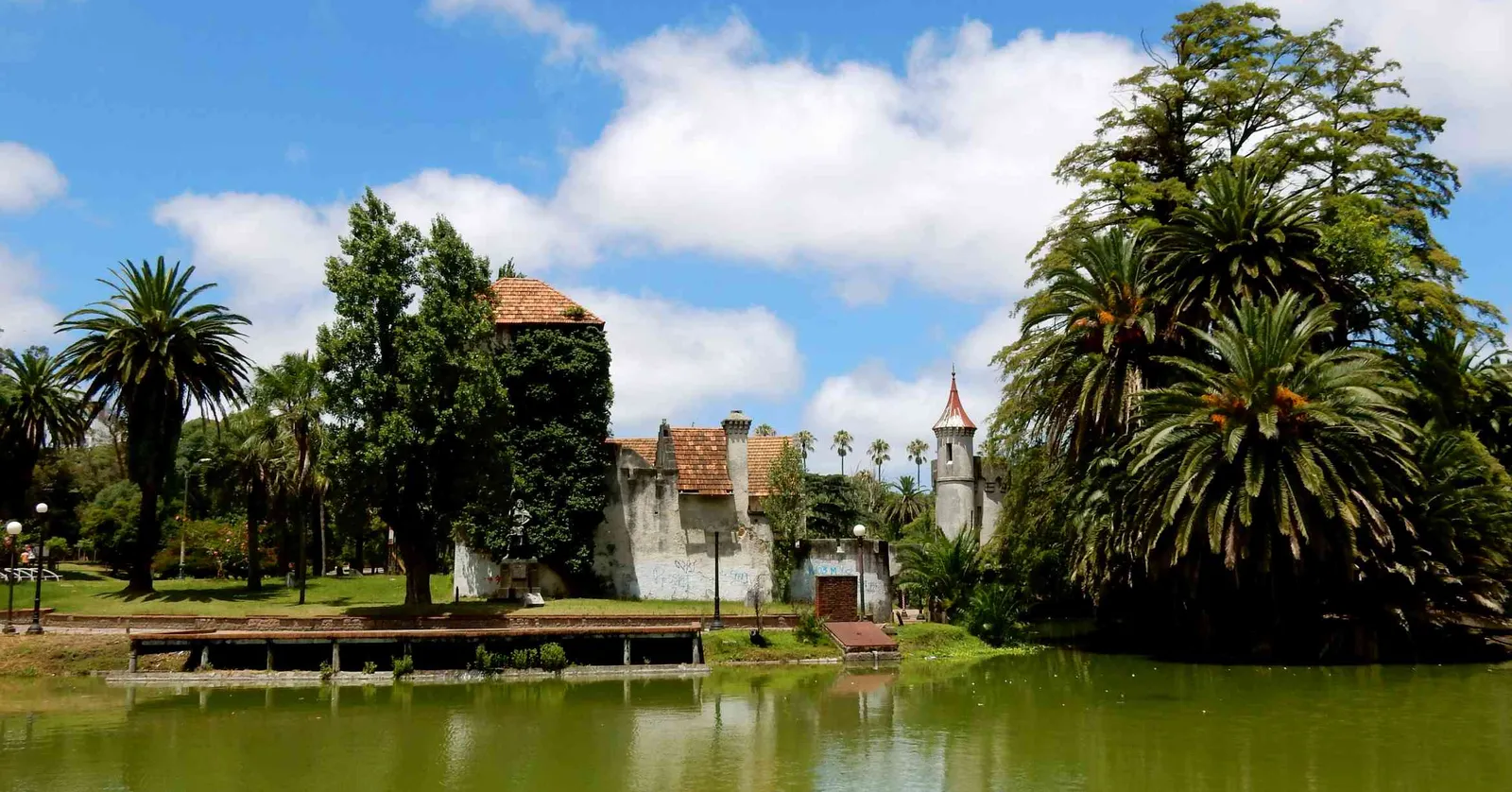 The Castillo del Parque Rodó in Montevideo, a mock-medieval stone castle with a turret and tiled roofs, reflected in the park's artificial lake and framed by palm trees under a blue sky