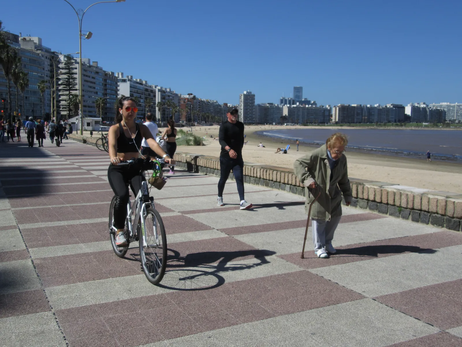 A cyclist and pedestrians on the checkerboard-paved Rambla in Pocitos, Montevideo, with the beach and city skyline running alongside on a clear day