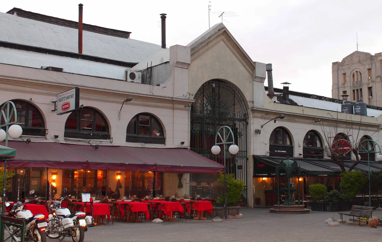 The arched arcade front of the Mercado del Puerto in Montevideo at dusk, lit from inside with red-clothed tables under an awning and Palacio Salvo's tower visible behind the rooftop